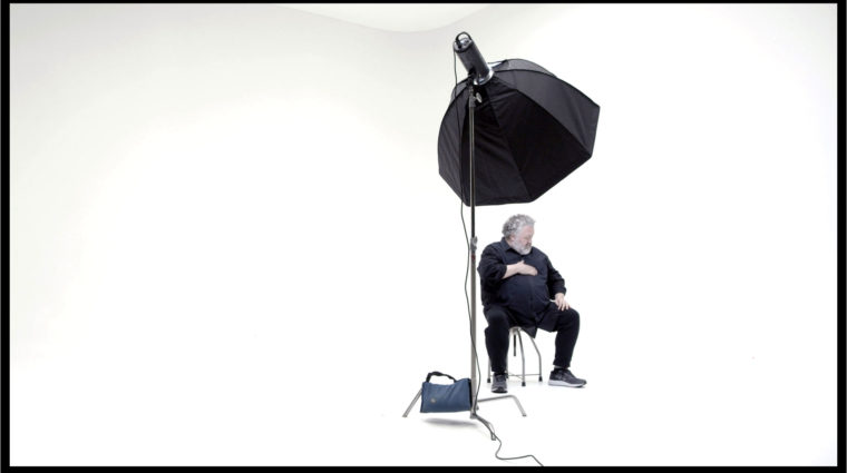 Bruce Mau sits under a film lamp in an empty, colorless room